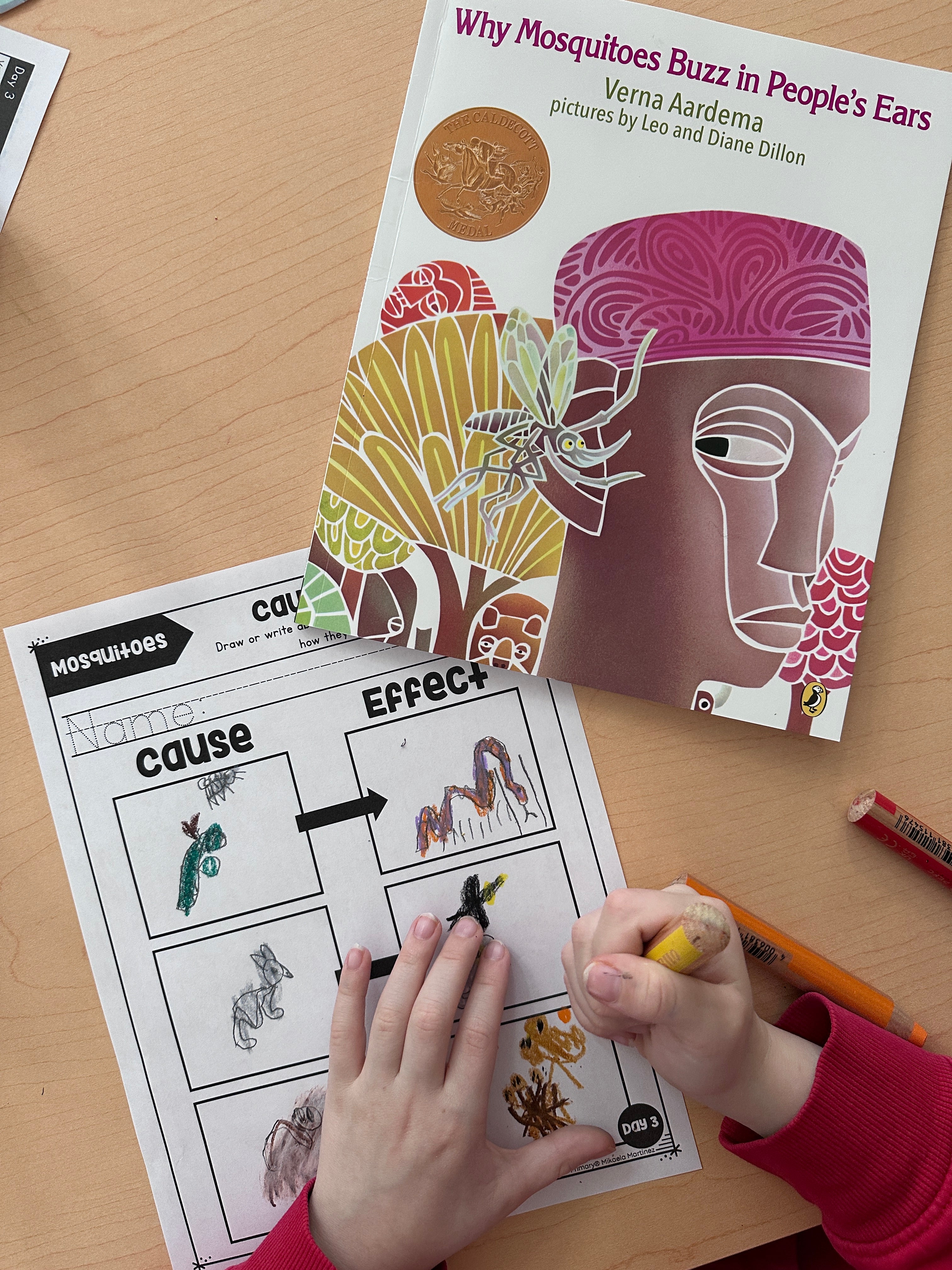 A child's hands are shown coloring a worksheet on a wooden desk, with the children's book 'Why Mosquitoes Buzz in People's Ears' by Diana Dellon visible underneath.