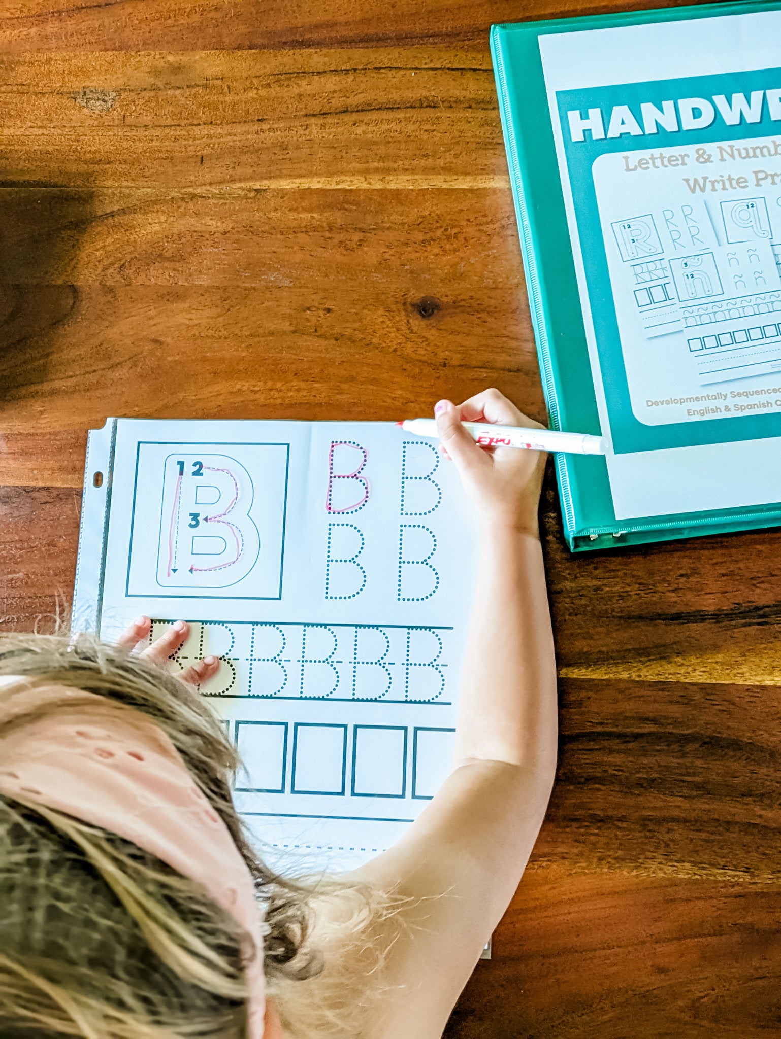 A child's hand and head are visible as they practice letter writing on a white worksheet with dotted letters and a pink marker, placed on a wooden table next to a teal binder labeled 'HANDWRITTEN'.