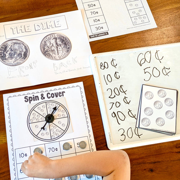 A child's hand interacting with a coin-themed educational activity sheet on a wooden table, surrounded by other matching worksheets and a coin storage tray.